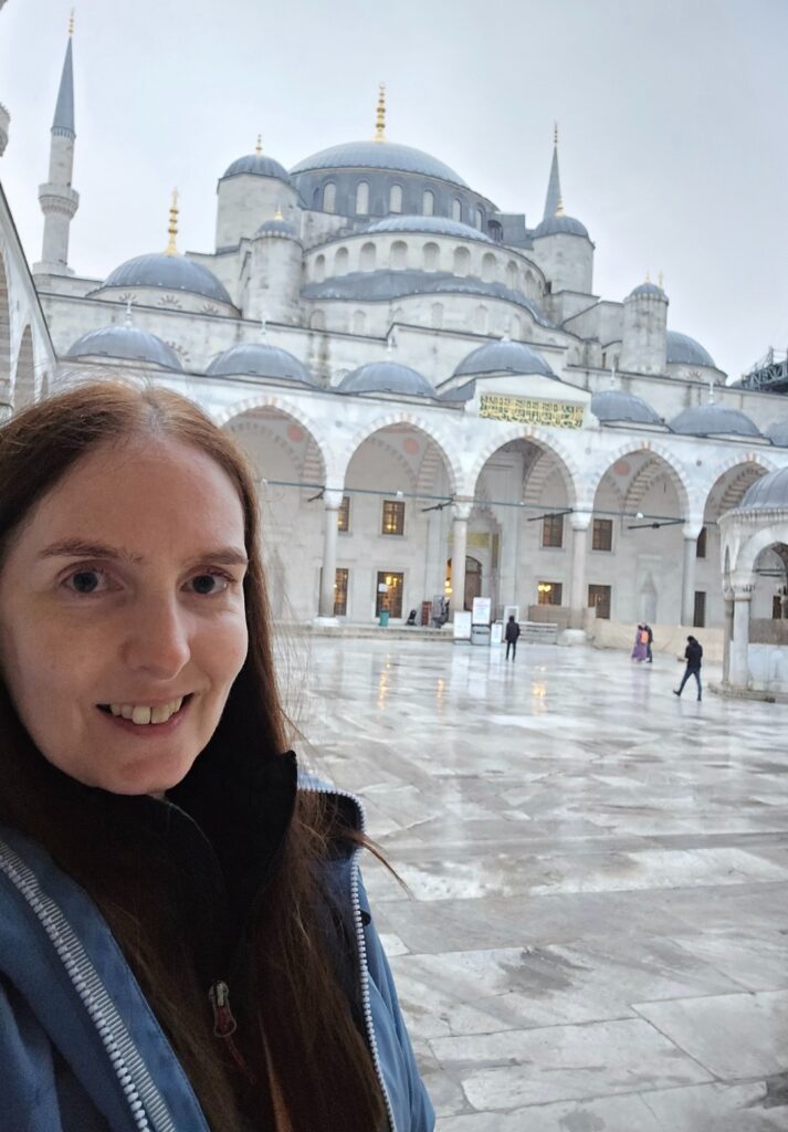 Me taking a selfie in the courtyard of the Blue Mosque on a cloudy day, with the grand domes and arched colonnade of the mosque rising behind her over a rain-slicked marble floor.