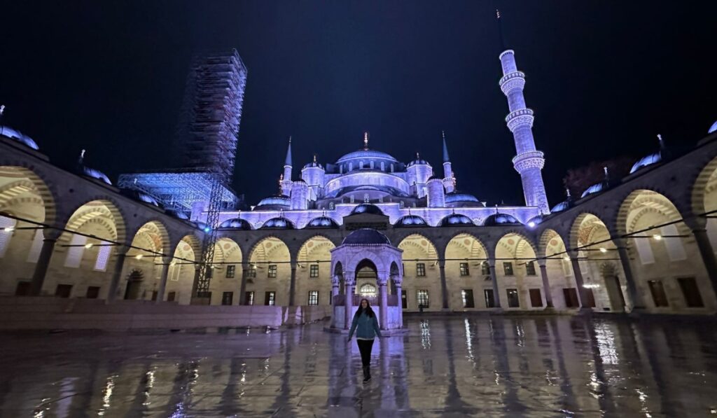 Me standing in the reflective, rain-slicked courtyard of the Blue Mosque at night, facing the camera as the mosque’s illuminated domes and arches frame the background under a dark sky.