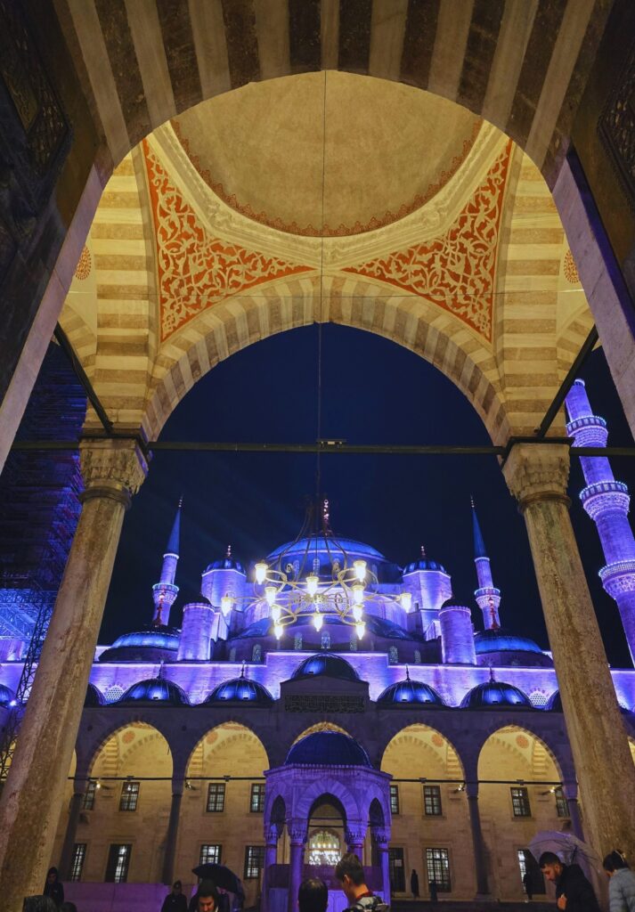 View of the illuminated Blue Mosque (Sultan Ahmed Mosque) in Istanbul at night, seen through a richly decorated archway with red and cream geometric patterns. A large chandelier hangs above the entrance courtyard where visitors gather under the lit domes and towering minarets.