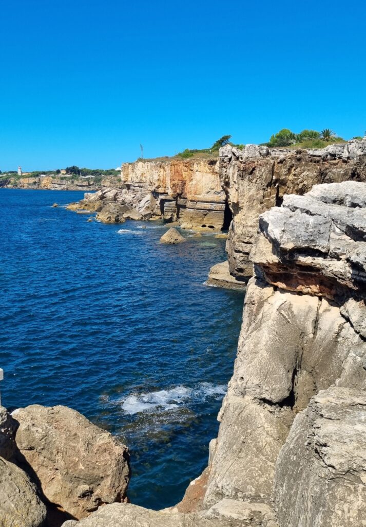 View of the dramatic cliffs at Boca do Inferno in Cascais, Portugal, with layered rock formations rising above the Atlantic Ocean under a bright blue sky.