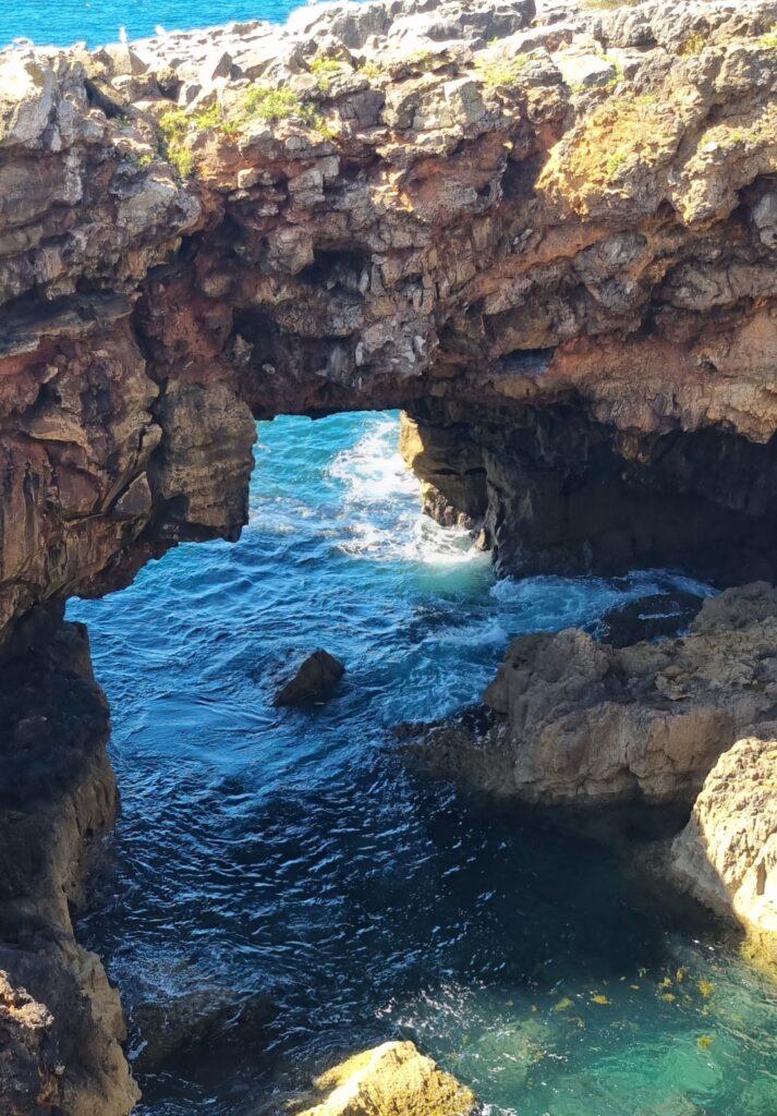 Natural sea arch at Boca do Inferno where waves crash through the rugged red and brown cliffside, creating a dramatic opening into the rock.