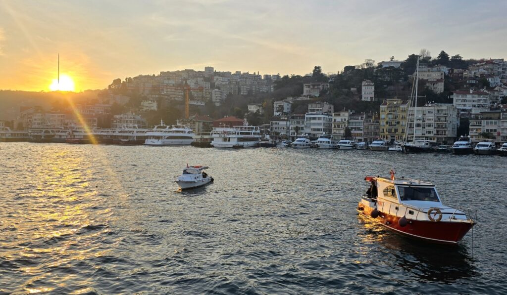 Small boats float on the Bosphorus at sunset near a marina, with a hillside densely packed with buildings and the last rays of sunlight hitting the rooftops and anchored yachts.