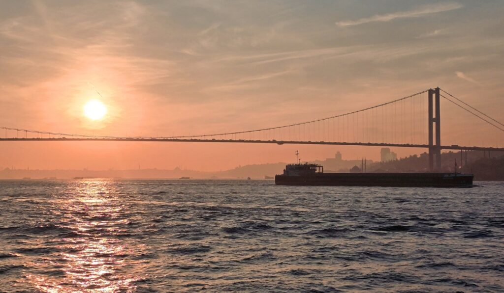 A cargo ship passes beneath the Bosphorus Bridge during sunset, with the pink-orange sky casting a hazy glow over the Istanbul skyline in the distance.
