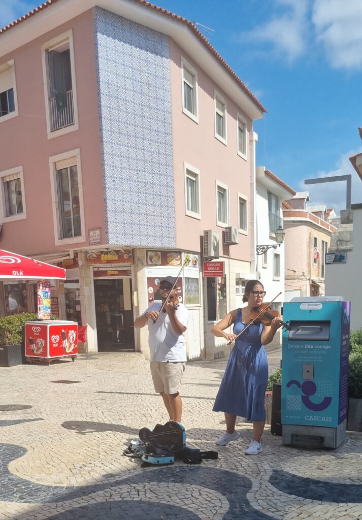 Two street musicians playing violins in a cobbled square in Cascais, standing in front of a pastel pink building and a kebab shop, with their violin case open for tips on the ground.