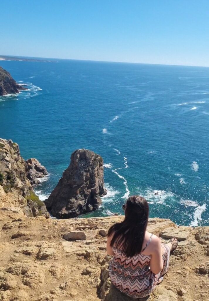 Me sitting on a rocky cliff edge overlooking the vast Atlantic Ocean at Cabo da Roca, with dramatic rock formations and deep blue water stretching into the horizon.