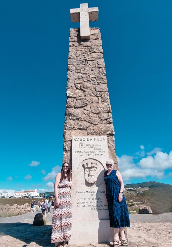 Me and my mum standing beside the stone monument at Cabo da Roca, marked with a cross and inscribed details identifying it as the westernmost point of mainland Europe, under a clear blue sky.