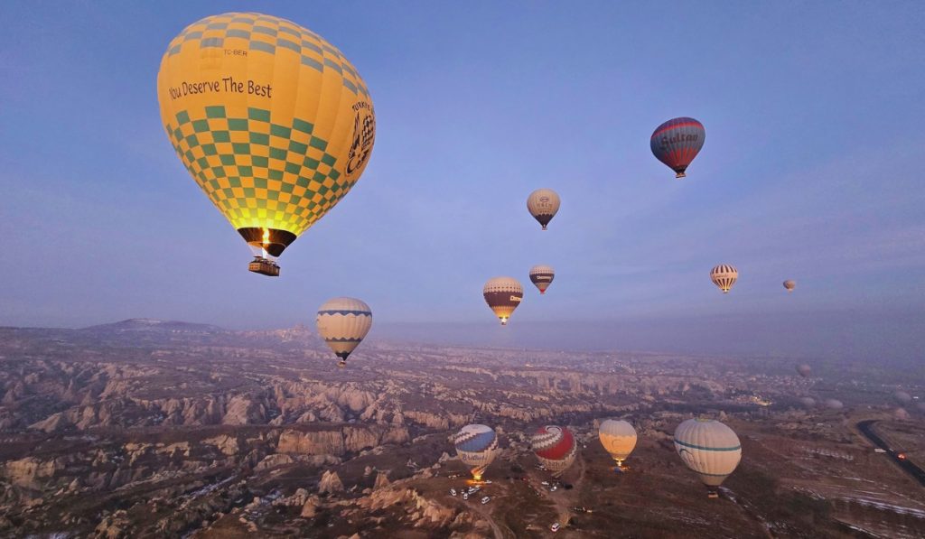 A cluster of colorful hot air balloons floats above the rocky valleys of Cappadocia, Turkey, at sunrise, with the prominent yellow-and-green balloon reading "You Deserve The Best." The early morning light casts a soft glow over the unique landscape of eroded cliffs and fairy chimneys below.