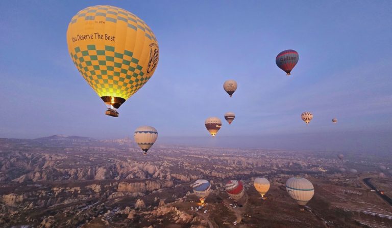 A cluster of colorful hot air balloons floats above the rocky valleys of Cappadocia, Turkey, at sunrise, with the prominent yellow-and-green balloon reading "You Deserve The Best." The early morning light casts a soft glow over the unique landscape of eroded cliffs and fairy chimneys below.
