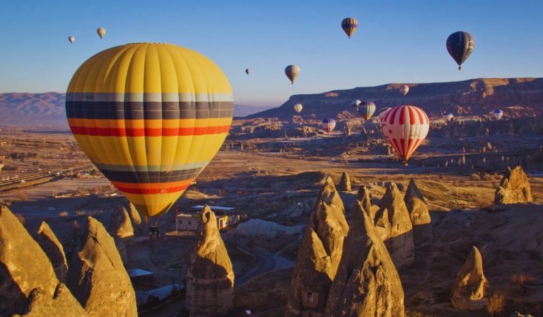 Multiple hot air balloons float above the unique rock formations of Cappadocia at sunrise, with the largest balloon in the foreground displaying bold yellow, red, and blue stripes. The landscape below is dotted with fairy chimneys and ridges, bathed in golden morning light.