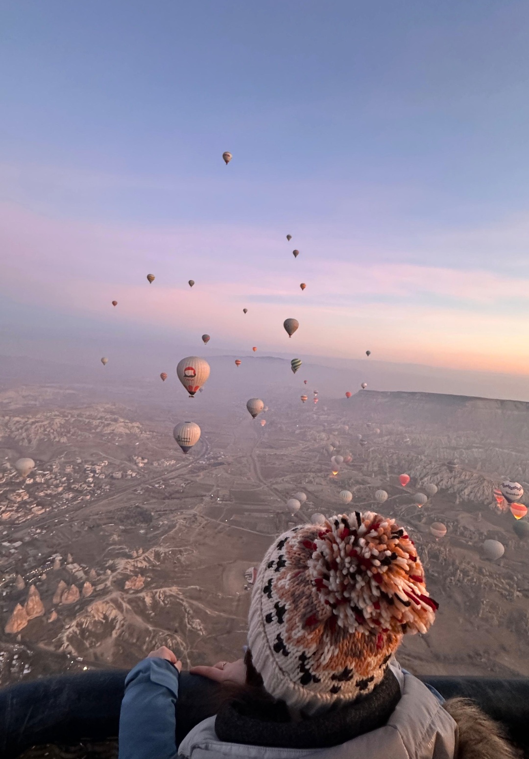 Me wearing a cozy hat and winter jacket looks out from a hot air balloon over Cappadocia at sunrise. Dozens of colorful balloons drift across the pastel sky and rugged, rocky landscape below, creating a dreamy, expansive view.