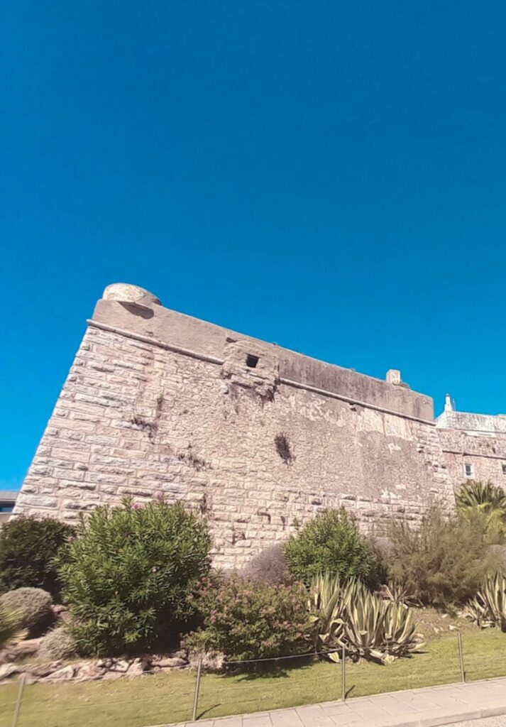 Exterior view of the Cascais Citadel, featuring massive stone ramparts with weathered textures and visible historic damage. The fortress rises above manicured greenery under a bright blue sky, showcasing its coastal defense heritage.