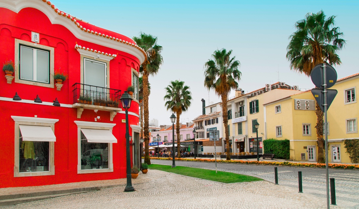 A vibrant street corner in Cascais with a striking red building on the left, lined with palm trees and pastel-colored buildings in the background. The red building features arched architectural details, window awnings, and flower pots, adding charm to the cobblestone pedestrian area.