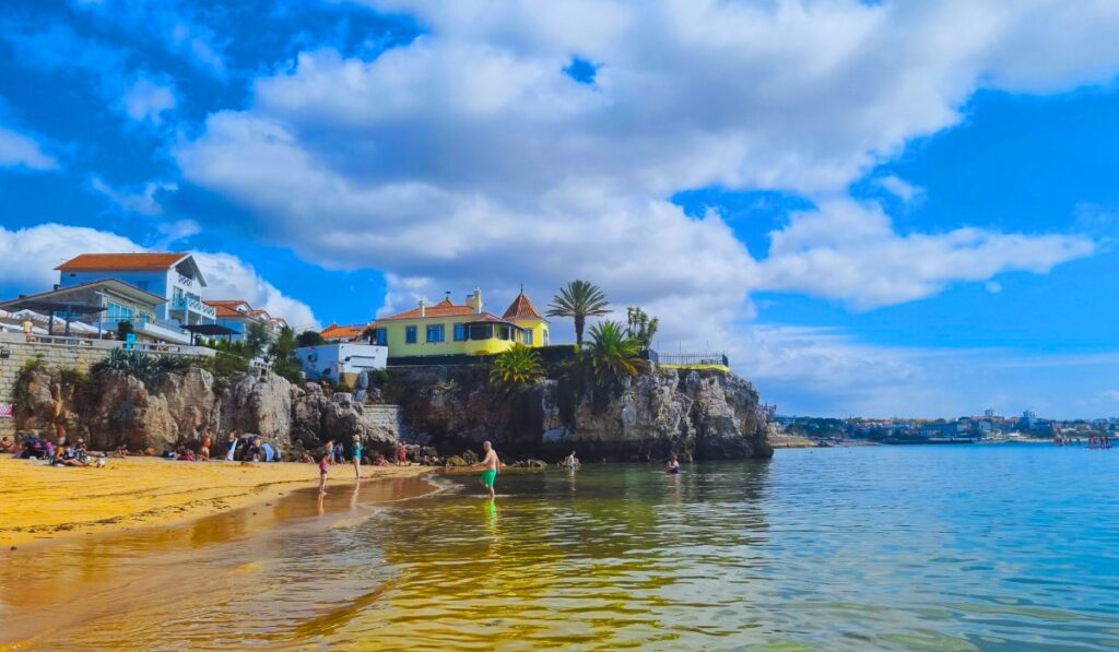 Praia da Rainha beach in Cascais with people relaxing and swimming near calm, clear water, backed by rocky cliffs and colorful houses including a standout yellow villa perched above, under a partly cloudy blue sky.