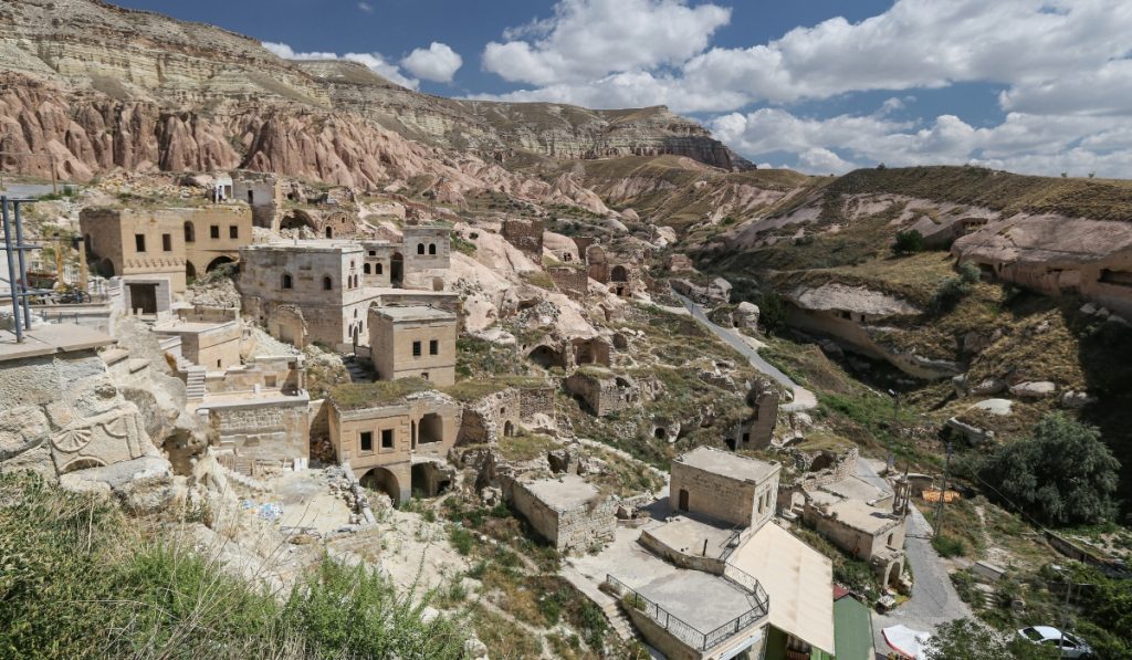 Panoramic view of an ancient village in Cappadocia, Turkey, with stone houses and cave dwellings built into the rocky hillside. The layered cliffs and arid terrain create a dramatic backdrop under a partly cloudy sky.