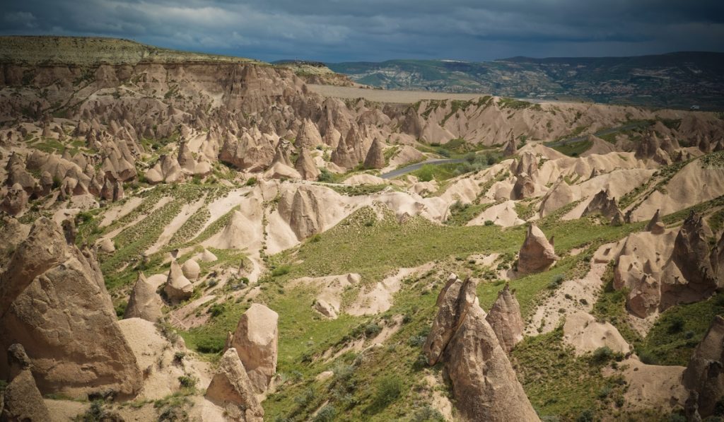 Expansive view of Cappadocia’s Devrent Valley, also known as Imagination Valley, featuring unusual rock formations and pointed fairy chimneys scattered across grassy slopes. The dramatic lighting under a moody sky highlights the surreal, almost lunar landscape.