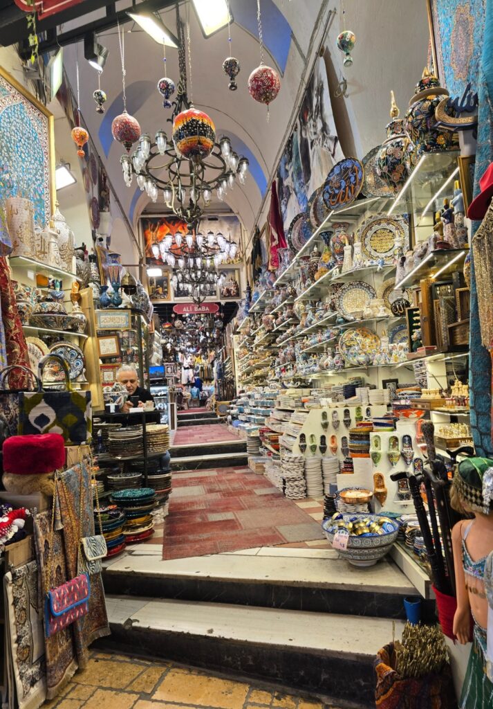 A narrow, chandelier-lit corridor in the Grand Bazaar packed with ceramic plates, bowls, decorative lamps, and Turkish souvenirs, with a shop sign reading “Ali Baba” visible in the distance.
