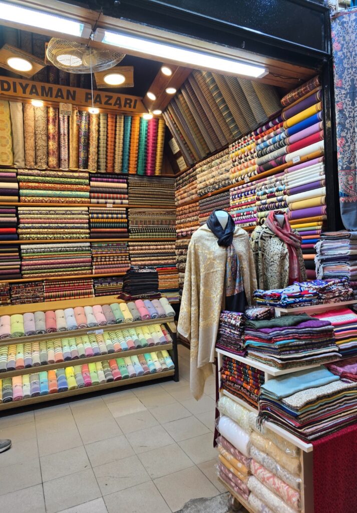 A fabric shop in the Grand Bazaar called “Adıyaman Pazarı” filled with neatly stacked and rolled textiles in vibrant colors and patterns, with two mannequins draped in traditional shawls and garments near the entrance.