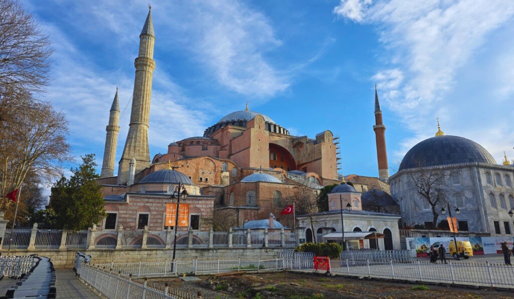 The iconic Hagia Sophia in Istanbul under a bright blue sky, showing its grand central dome, surrounding smaller domes, and four tall minarets. The foreground includes the perimeter fence, Turkish flags, and nearby smaller domed buildings within the complex.