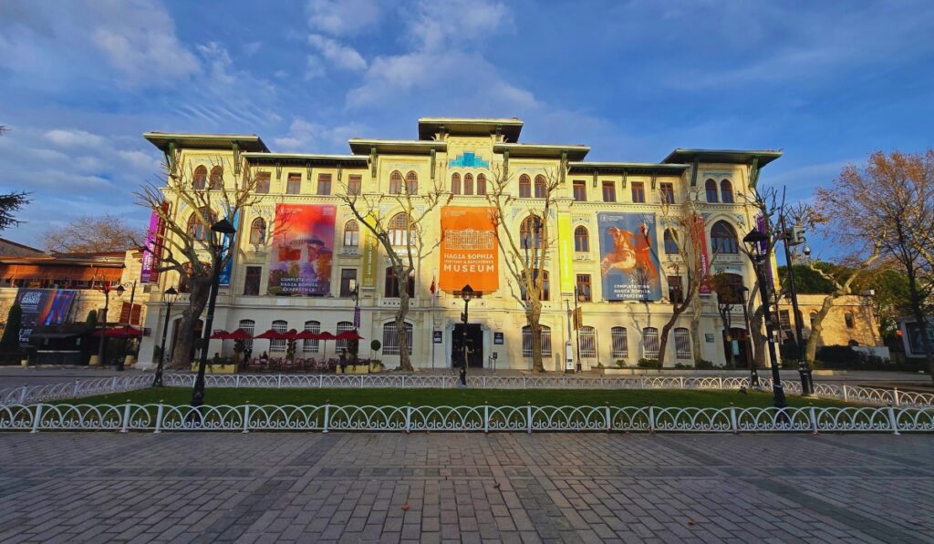 Front view of the Hagia Sophia History and Experience Museum in Istanbul, a pale yellow historic building with tall windows and colorful banners advertising exhibitions. Leafless trees and ornate white fencing line the cobblestone square in the warm light of late afternoon.