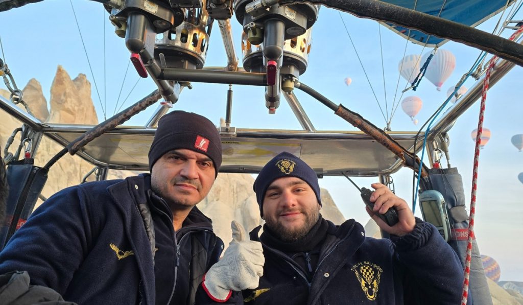 Two hot air balloon pilots stand inside the balloon basket, dressed warmly in navy uniforms and beanies, with one giving a thumbs-up and the other holding a radio. Behind them, numerous balloons float over the rock formations of Cappadocia, Turkey, under a clear morning sky.
