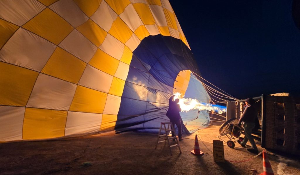 A hot air balloon is being inflated in the early morning darkness, with one person standing on a ladder managing the flame burner as fire shoots into the balloon’s colorful yellow, white, and blue envelope. Another person operates a fan to help fill the balloon with air, while caution cones and a sign reading "Do not cross this line" mark the safety zone.