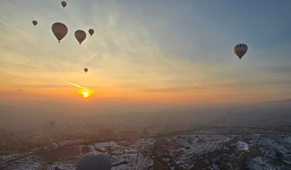 Hot air balloons drift across the sky at sunrise over a misty, snow-dusted landscape in Cappadocia, Turkey. The glowing sun casts a golden hue across the horizon, silhouetting the balloons against the soft gradient of morning light.