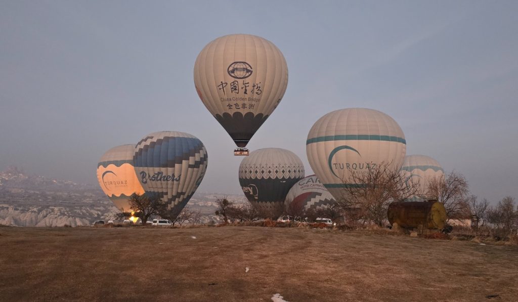 Several hot air balloons prepare for takeoff at dawn in Cappadocia, Turkey, with one balloon already airborne and others inflating on the ground. Each balloon displays branding, including “China Golden Bridge,” “Brothers,” and “TURQUAZ,” set against a backdrop of rocky valleys and a hazy sky.