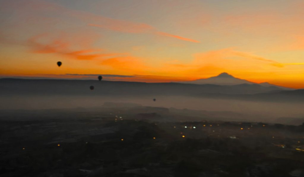 A hazy sunrise view over Cappadocia with a silhouette of Mount Erciyes in the distance, its peak bathed in soft orange and pink light. A few hot air balloons drift through the misty sky above the faintly glowing lights of the town below.