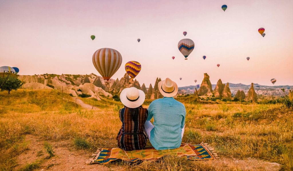 A couple wearing straw hats sits on a colorful picnic blanket in a golden field, watching dozens of hot air balloons float above the rocky fairy chimneys of Cappadocia during sunrise. The sky glows with warm pastel hues, creating a romantic and dreamy atmosphere.