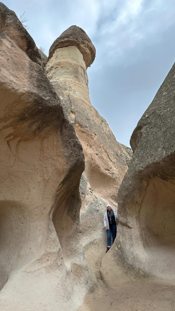 Me in a winter jacket and beanie stands between towering, smooth rock formations in Cappadocia, framed by curving volcanic walls beneath a cloudy sky. One large fairy chimney with a dark cap rises behind her, showcasing the region’s dramatic natural geology.