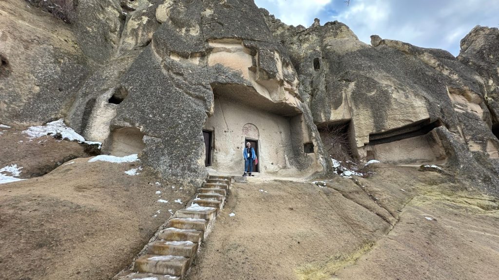 Standing at the entrance of a rock-carved church in Cappadocia, reached by a narrow staircase cut into the soft stone hillside. The cliff face features multiple small openings and a faded red fresco above the arched doorway, hinting at the site's ancient Christian history.