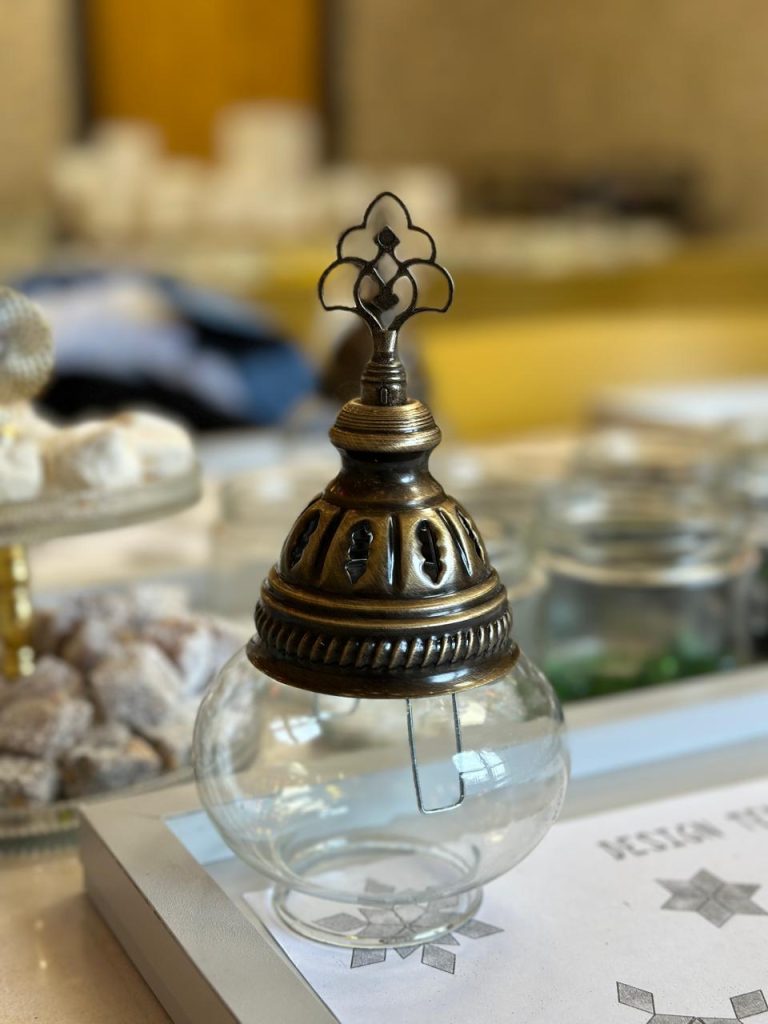 Close-up of a clear glass lamp base with an ornate bronze-colored top, resting on a design template during a mosaic lamp-making workshop. Small jars of colored beads and pieces are visible in the background.