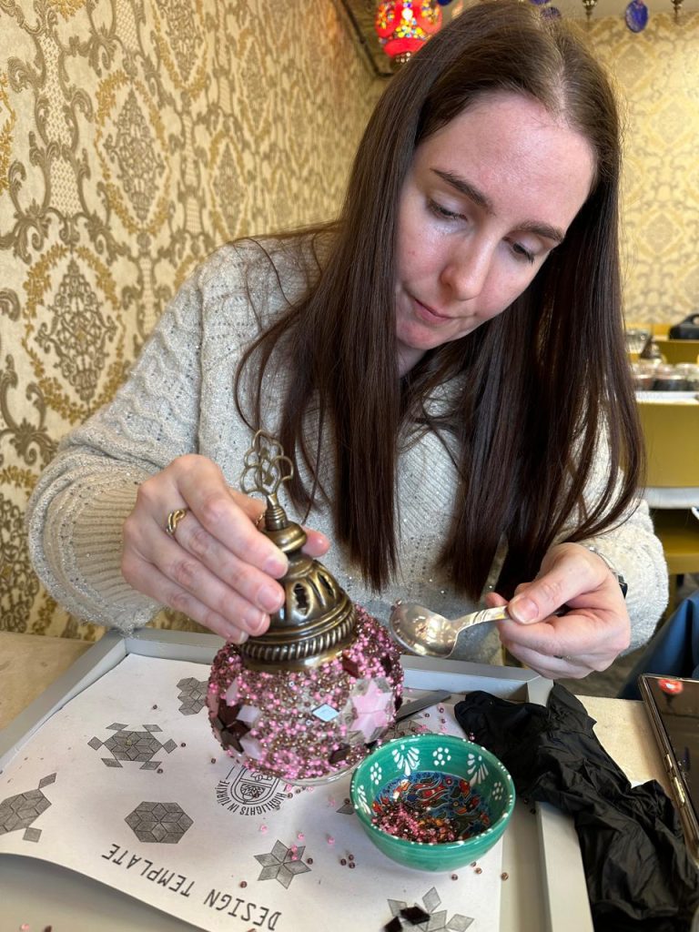I am concentrating as I add pink and gold beads to a mosaic lamp using a spoon, carefully decorating the glass surface. The lamp rests on a patterned template tray beside a small dish of beads in a Turkish mosaic workshop setting.