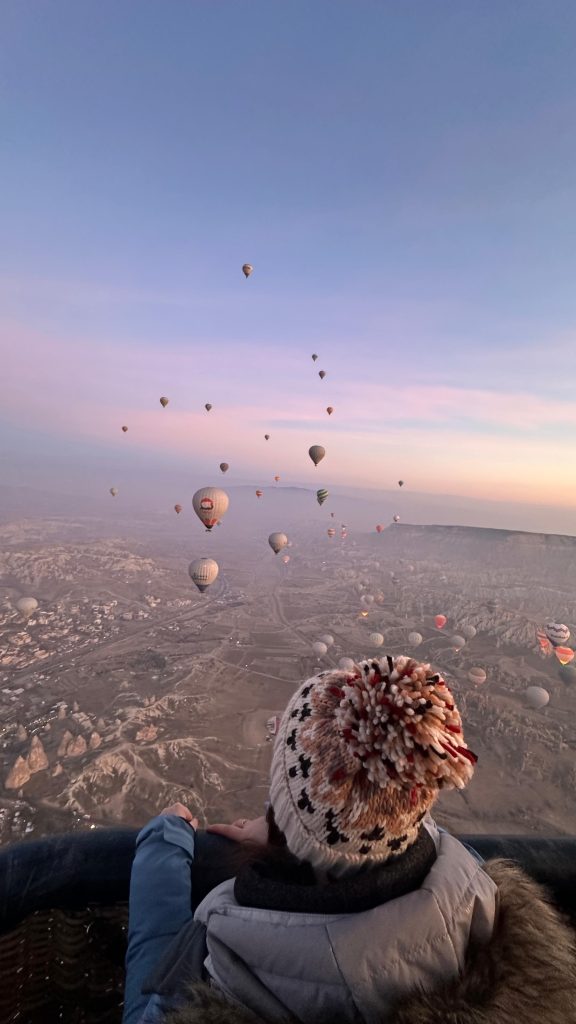 View from a hot air balloon high above Cappadocia at sunrise, with dozens of colorful balloons floating over the rocky terrain. A person wearing a winter hat with a pom-pom leans over the edge of the basket, taking in the expansive landscape.
