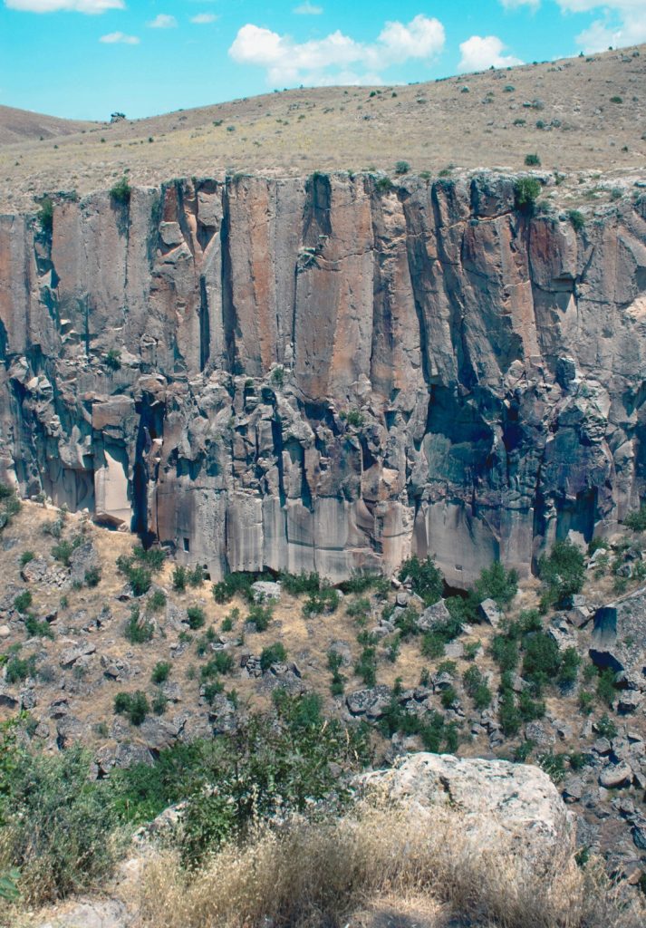 Steep, rugged cliffs of Ihlara Valley in Cappadocia, featuring vertical rock walls with subtle reddish tones and scattered greenery along the canyon floor. The dramatic geological formations rise from the dry, rocky landscape under a bright blue sky.