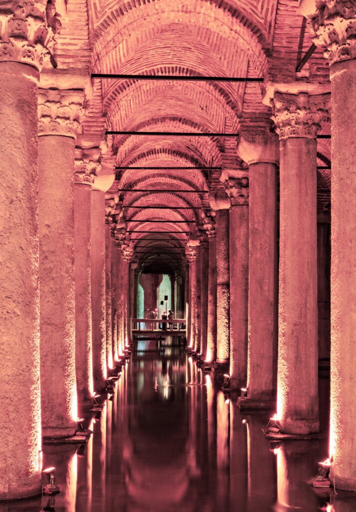 Central corridor of the Basilica Cistern illuminated in pink lighting, with tall stone columns and barrel-vaulted brick ceilings stretching into the distance. Visitors stand at the far end on a wooden platform, surrounded by the still, reflective water below.
