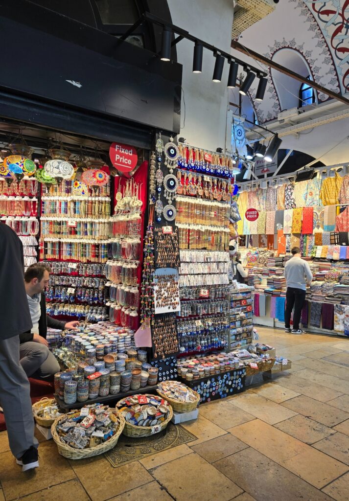 Colorful souvenir stall in the Grand Bazaar offering evil eye charms, keychains, trinkets, bracelets, and textiles, with red signs marking “Fixed Price” and baskets of small items displayed on the floor.