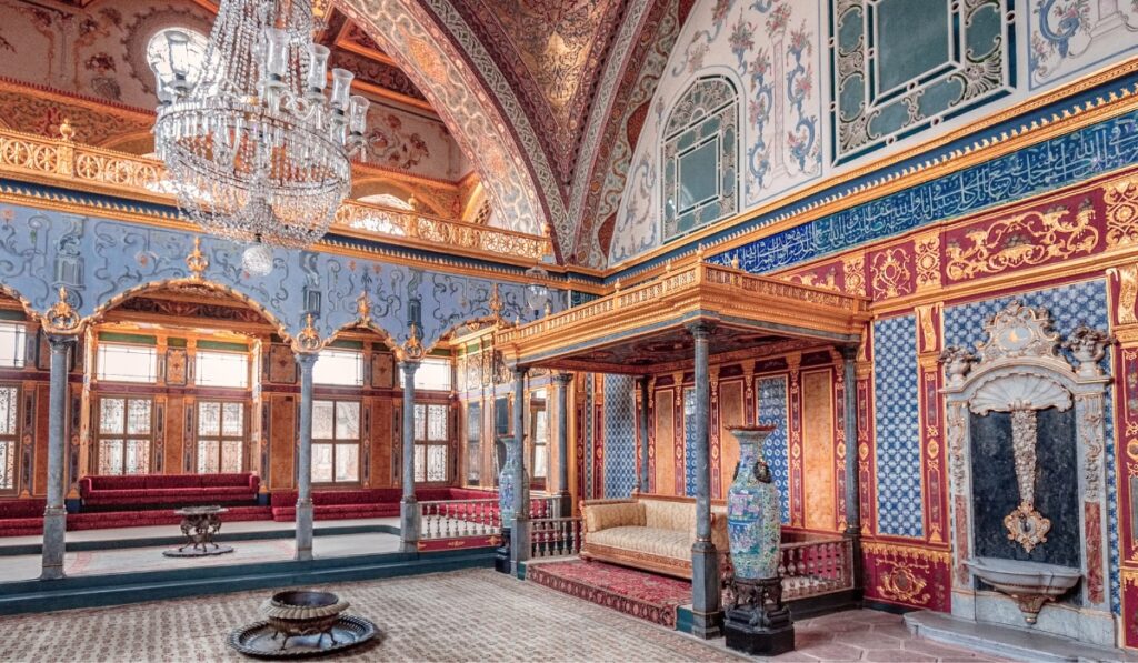 Lavishly decorated interior room of Topkapı Palace featuring intricate tilework, gold accents, a large crystal chandelier, and an ornate wooden canopy bed. Red velvet seating lines the windows under blue and gold arches, with a marble fountain and tall decorative vases enhancing the opulent Ottoman design.