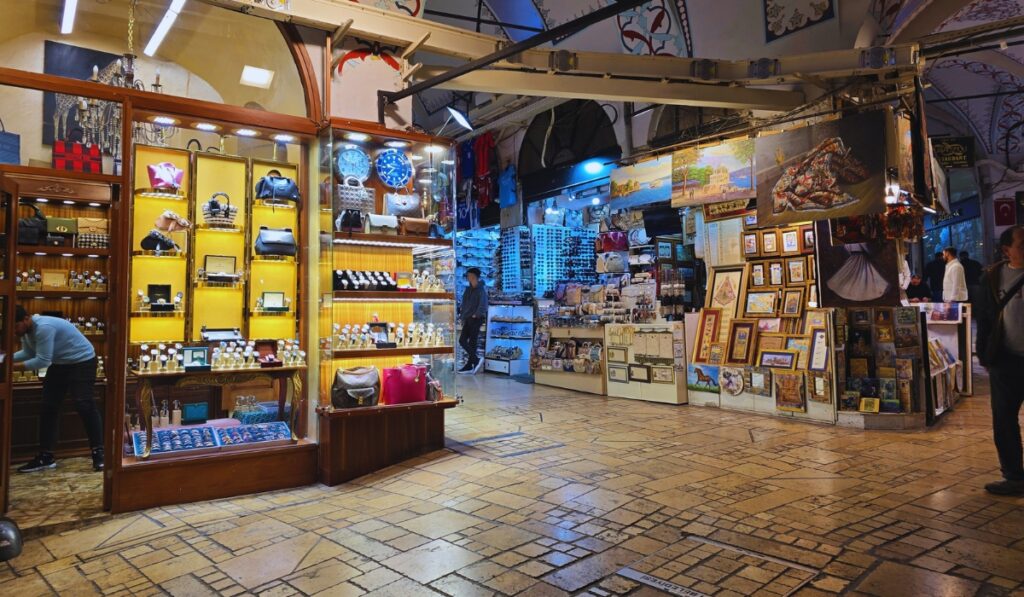 Interior of Istanbul’s Grand Bazaar showing a jewelry and leather goods shop with brightly lit displays of watches, handbags, and accessories, next to a stall selling paintings, souvenirs, and framed artwork under the arched, decorated ceiling.