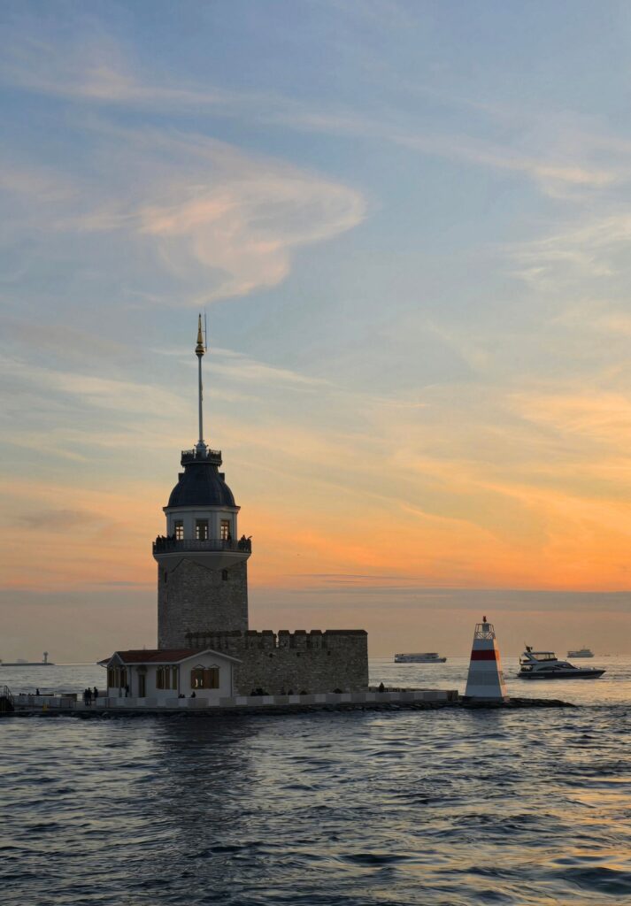 The Maiden’s Tower (Kız Kulesi) stands on a small islet in the Bosphorus at dusk, its lighthouse softly illuminated as boats pass in the background under a pastel-colored sky.