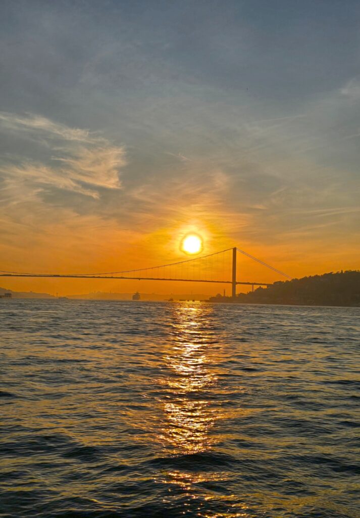 The sun sets over the Bosphorus Strait, casting golden reflections on the water with the silhouette of the Bosphorus Bridge stretching across the horizon beneath a glowing orange sky.