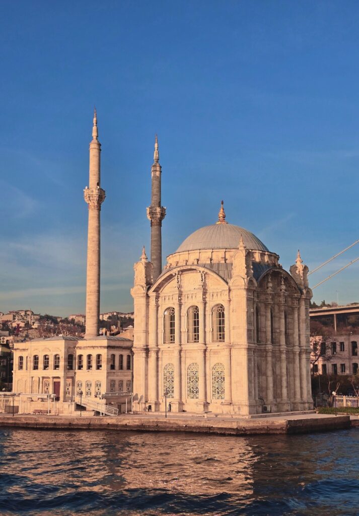 Ortaköy Mosque shines in warm golden light beside the Bosphorus, with its twin minarets and ornate Baroque-style architecture standing out against a clear blue evening sky.


Sixth Image: A woman stands at the back of a boat cruising along the Bosphorus, gazing toward the Bosphorus Bridge and a red ship as the sun sets behind the hills in the distance.