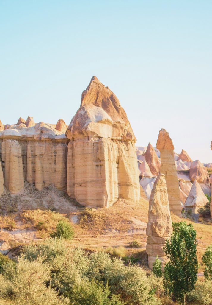 Sunlit view of fairy chimneys in Cappadocia, featuring a large, sharply pointed rock formation surrounded by smooth, pillar-like structures. The soft golden light enhances the warm tones of the eroded volcanic landscape.