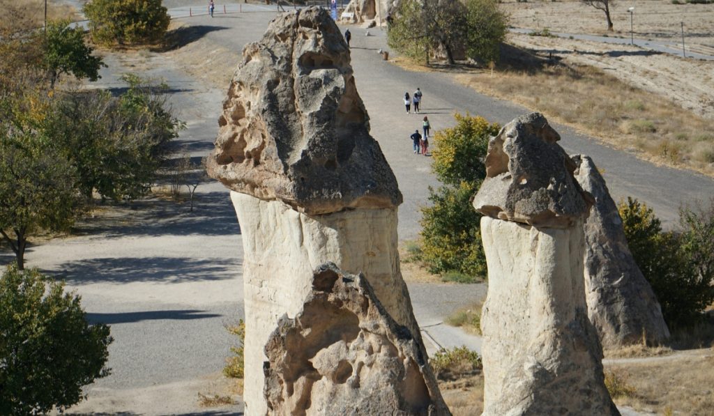 Close-up of two prominent fairy chimneys with dark mushroom-like caps, located near a paved walking path where visitors stroll through the landscape. The perspective emphasizes the unique shape and size of the formations.
