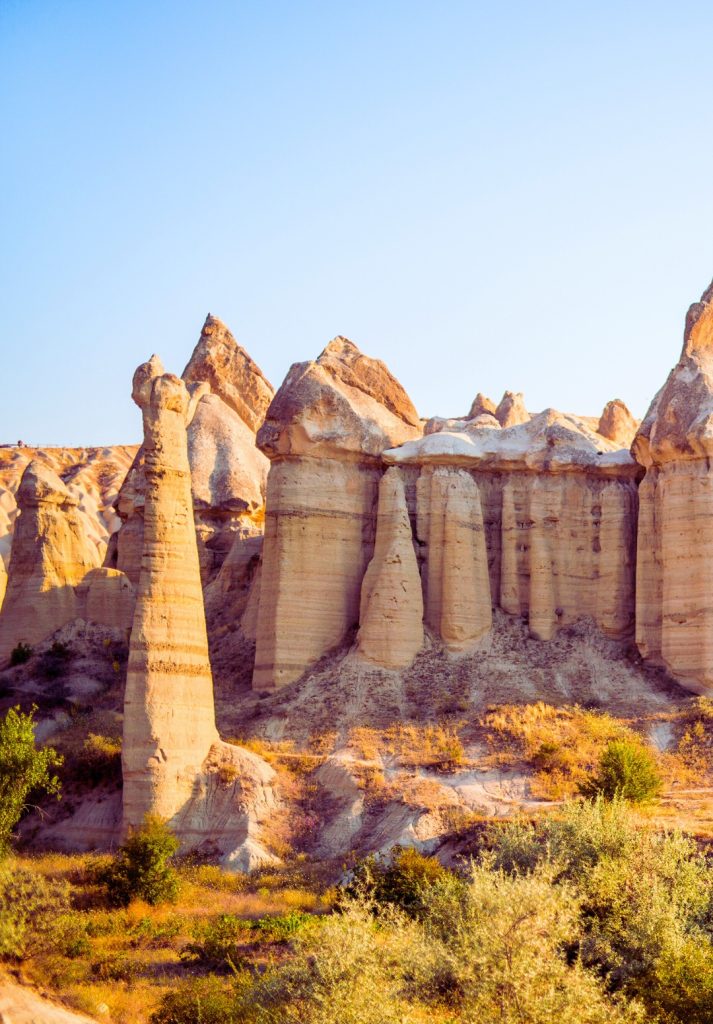 Tall and narrow fairy chimneys stand closely grouped in Cappadocia under a clear blue sky. The formations show vertical striations and dark caps, highlighting their wind-carved textures.
