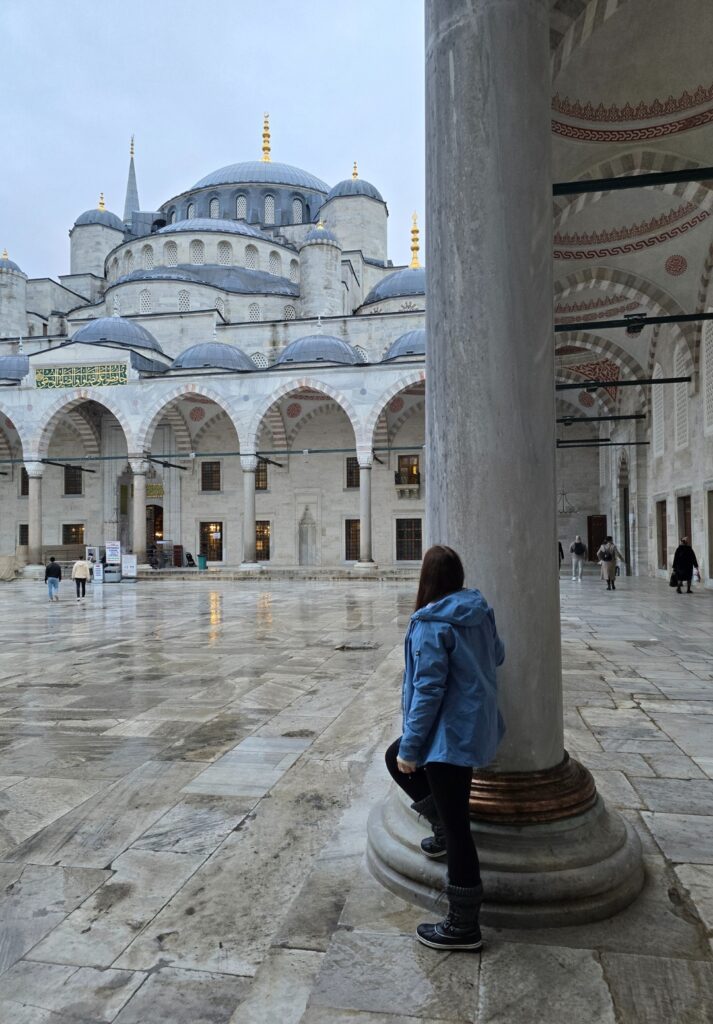 Me in a blue jacket leans against a marble column in the Blue Mosque’s courtyard, looking toward the mosque’s domes and arches as other visitors walk across the wet stone floor on a grey, rainy day.