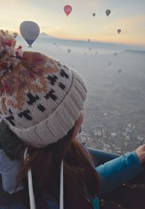 A woman in a cozy knit hat with a pom-pom looks out from a hot air balloon basket, watching dozens of colorful balloons float above the misty landscape of Cappadocia at sunrise. The soft light highlights the mountainous horizon and the scattered town below.