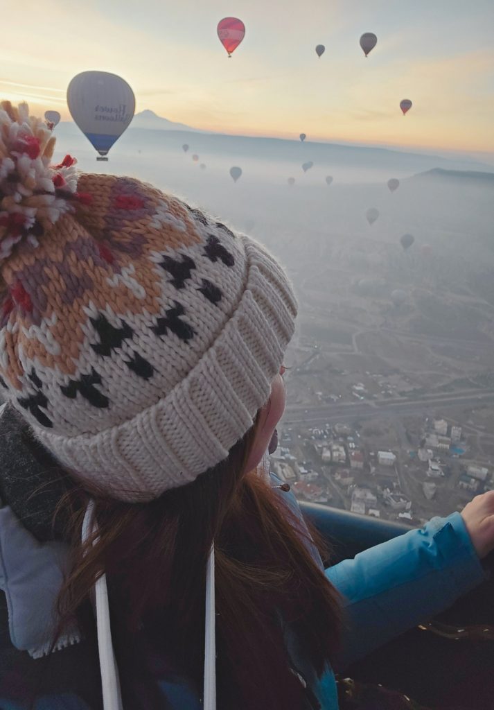 A woman in a cozy knit hat with a pom-pom looks out from a hot air balloon basket, watching dozens of colorful balloons float above the misty landscape of Cappadocia at sunrise. The soft light highlights the mountainous horizon and the scattered town below.