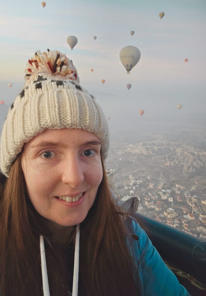 A woman wearing a cream-colored knit hat with a pom-pom takes a selfie during a hot air balloon ride, with dozens of colorful balloons floating in the misty sky over Cappadocia’s rocky terrain and villages in the background.
