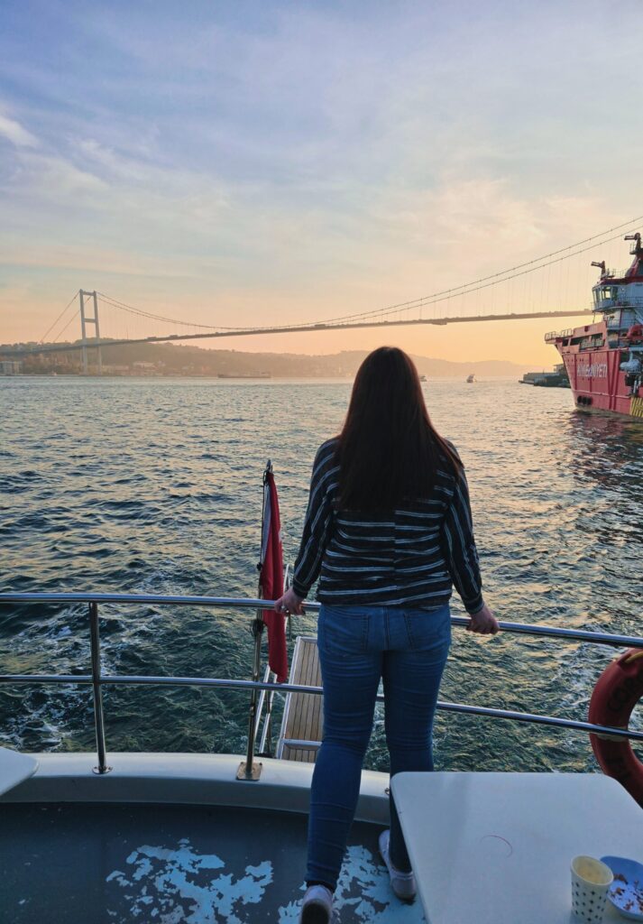 Me standing at the back of a boat cruising along the Bosphorus, gazing toward the Bosphorus Bridge and a red ship as the sun sets behind the hills in the distance.